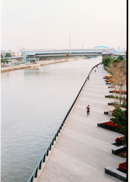 Free Tranquil riverside view with walking path, bridge, and minimal human activity. Stock Photo