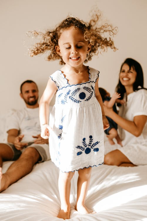 Adorable little girl with curly hair in light dress jumping on bed with closed eyes near happy young parents and newborn baby