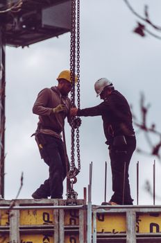Two workers in safety gear operate a chain hoist at a construction site.