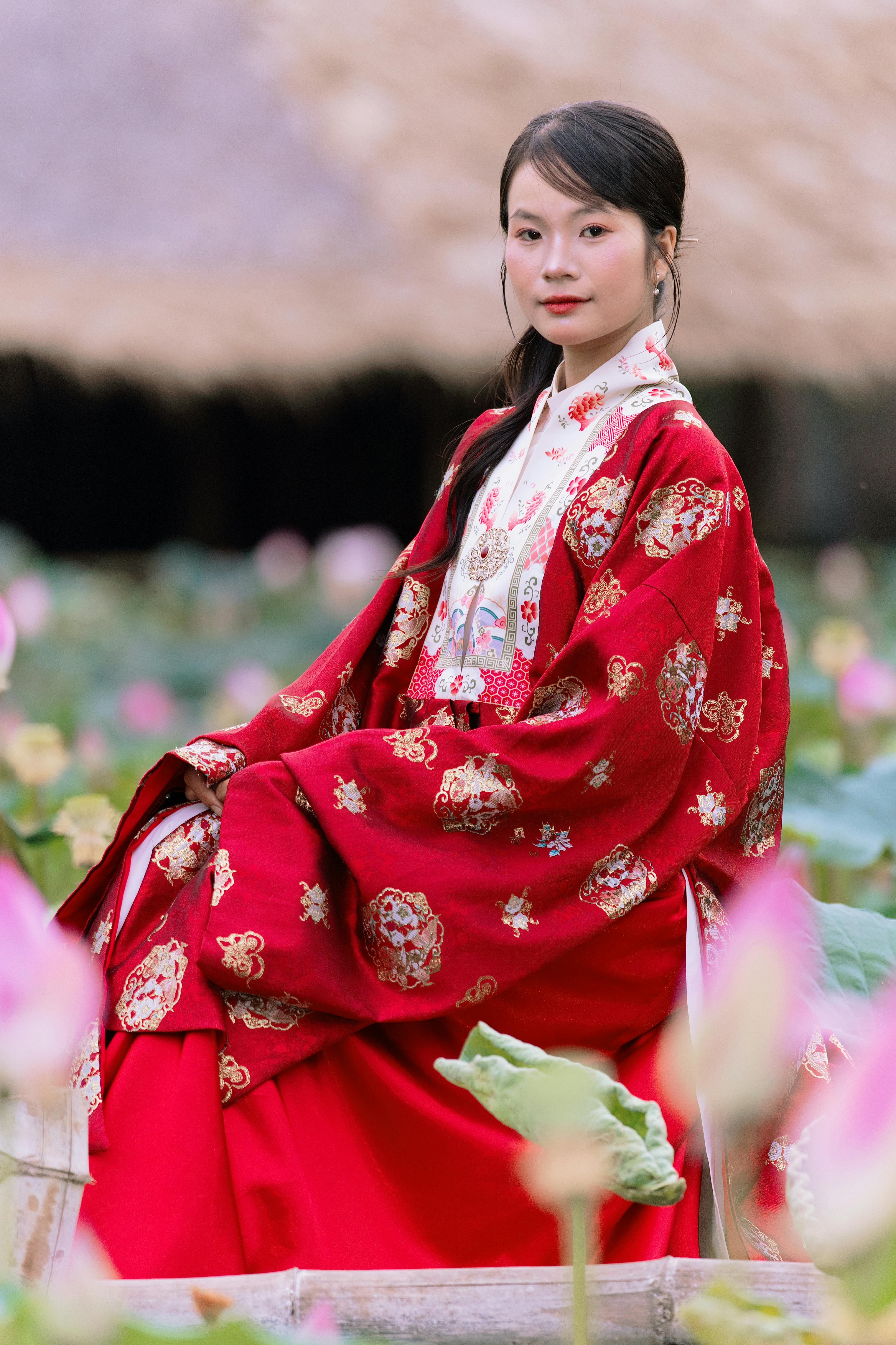 Woman in red traditional dress seated among blooming lotus, captured in serene setting.