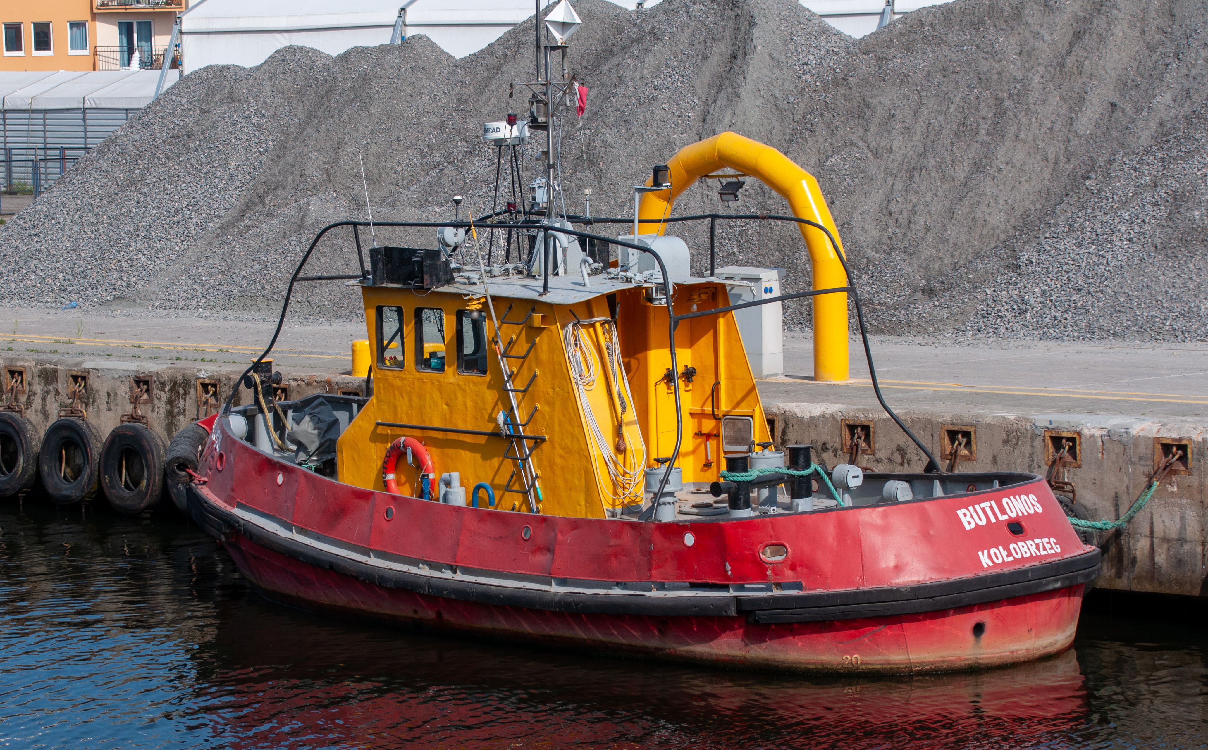 Colorful red and yellow tugboat docked at a harbor, surrounded by industrial materials.