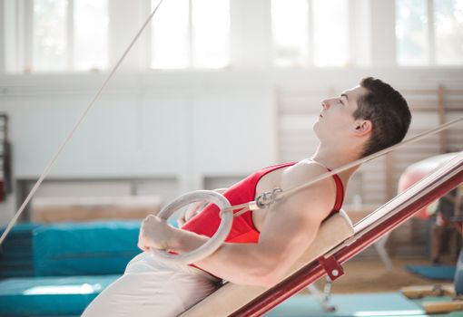 Side view of young sportsman in activewear doing exercise while lying on professional sport equipment with rings