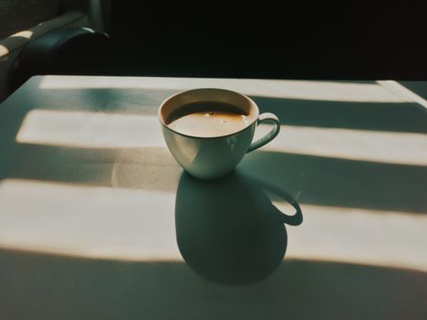 A white coffee cup sits on a table with striking sunlight and shadows creating a warm ambiance.
