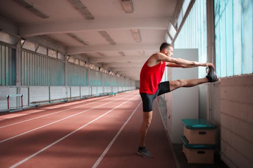 Zijaanzichtfoto Van De Mens In Rode Tanktop En Zwarte Korte Broek Die Zich Uitstrekt In Indoor Atletiekbaan