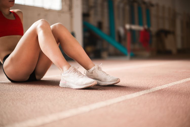 Photo Of Woman In Red Tank Top, Black Shorts, And White Sneakers Sitting On Running Track