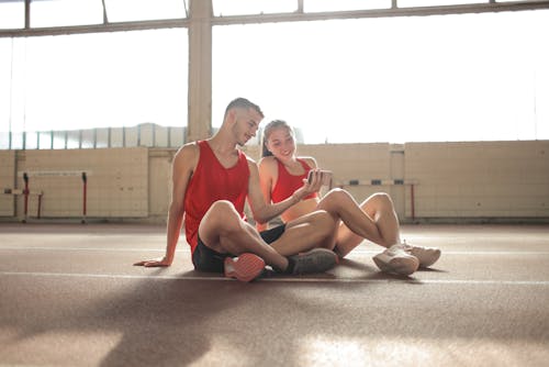 Happy young sportsmen sitting with crossed legs on floor and surfing smartphone together at sports hall