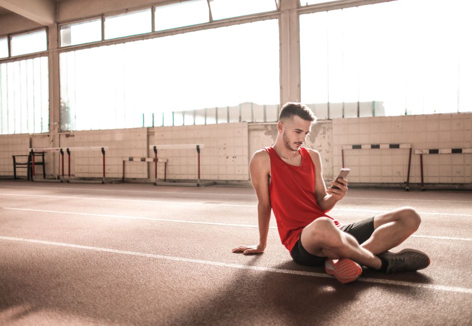 Young male athlete in red sportswear sitting on indoor track using smartphone, embracing modern technology.
