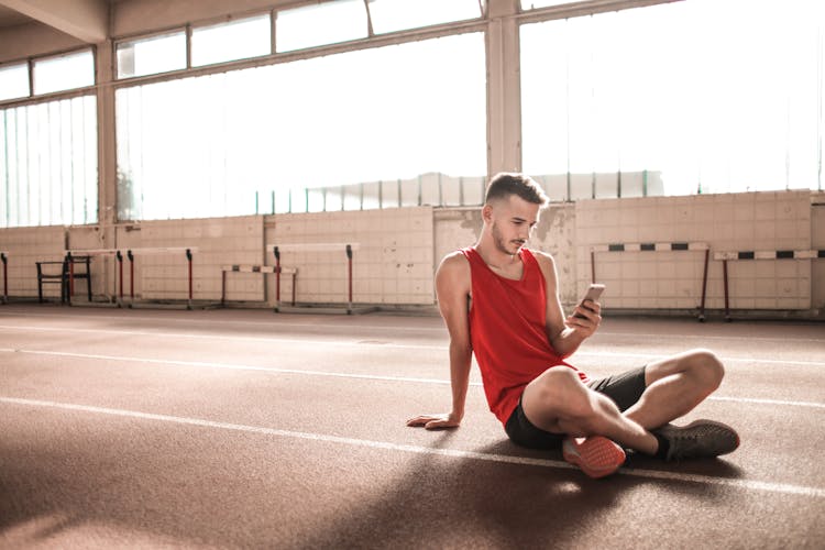 Man In Red Tank Top And Black Shorts Sitting On Running Track While Texting