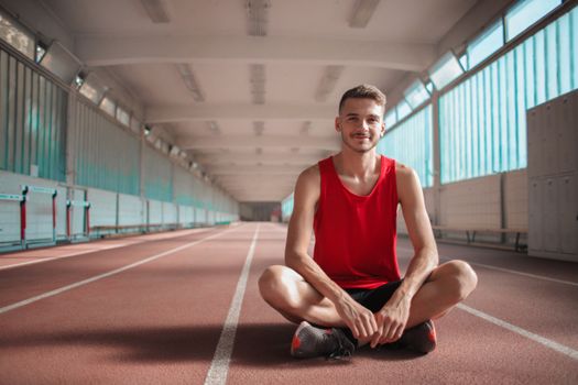 A happy male athlete in a red shirt sitting on an indoor track, relaxing after exercise.