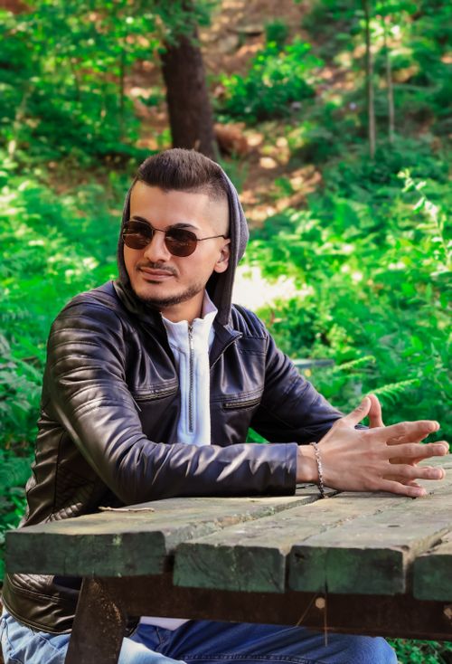 Free Man in leather jacket and sunglasses sitting at a wooden table in a lush green forest. Stock Photo
