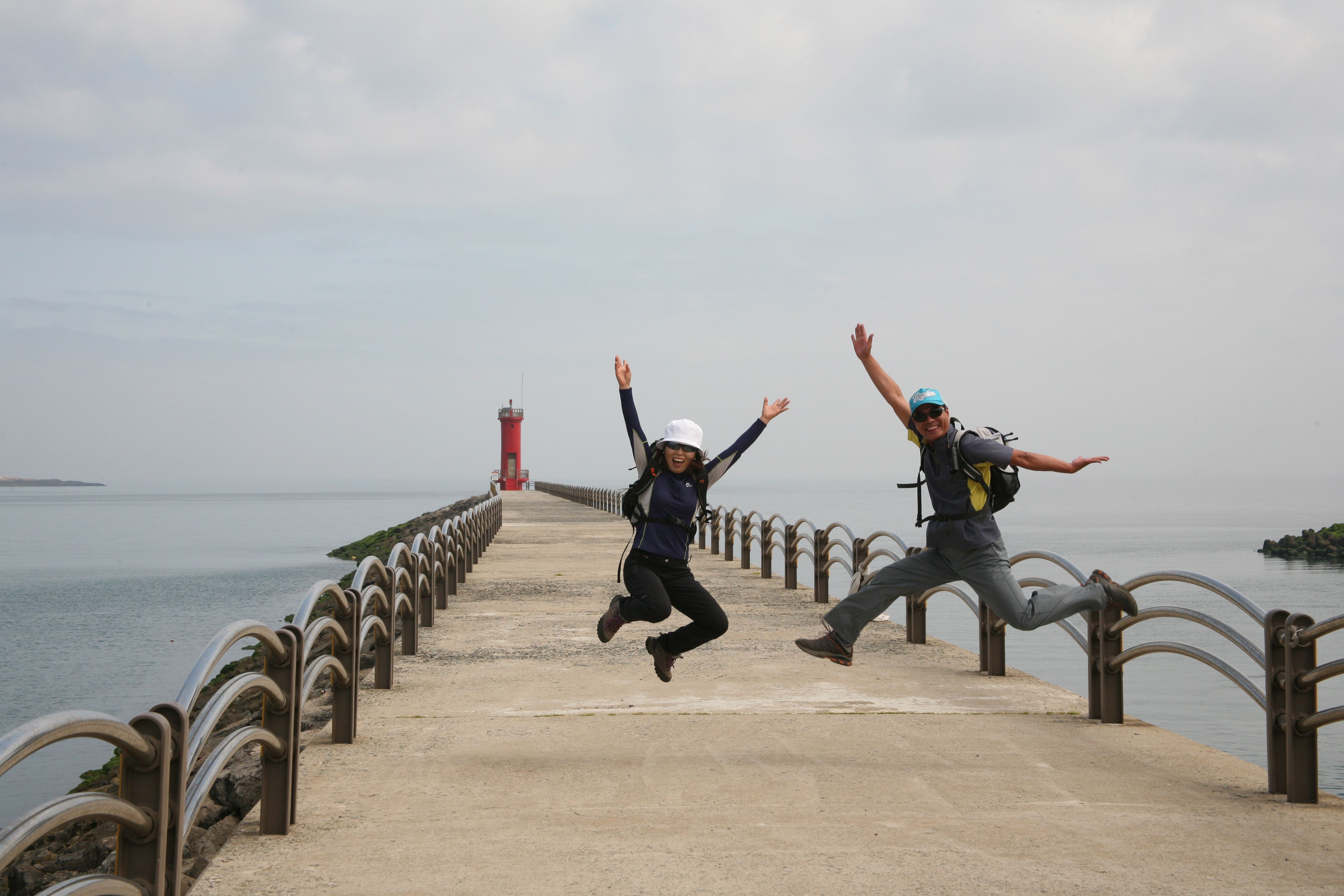Two adults jump in joy on a pier near a red lighthouse with ocean backdrop.