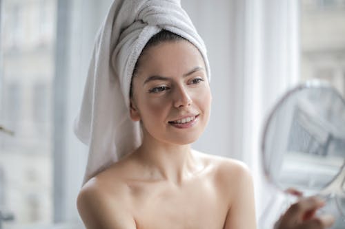Selective Focus Portrait Photo of Smiling Woman With a Towel on Head Looking in the Mirror 