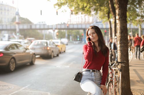 Selective Focus Photo of Woman in Red Long Sleeve Sweater and Blue Jeans Sitting on Metal Railing  While Talking on the Phone