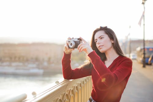 Free Selective Focus Photo of Woman in Red Sweater Taking a Picture with a Black Camera While Standing by Bridge Railing Stock Photo