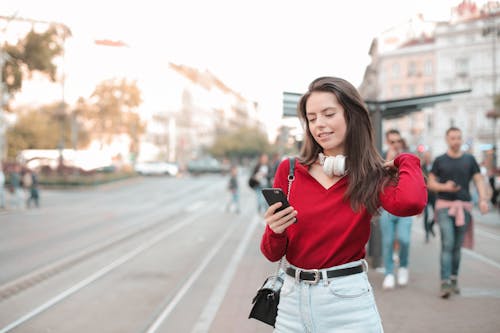 Selective Focus Photo of Woman in Red Sweater and Blue Denim Jeans Using Her Phone While Standing on Sidewalk