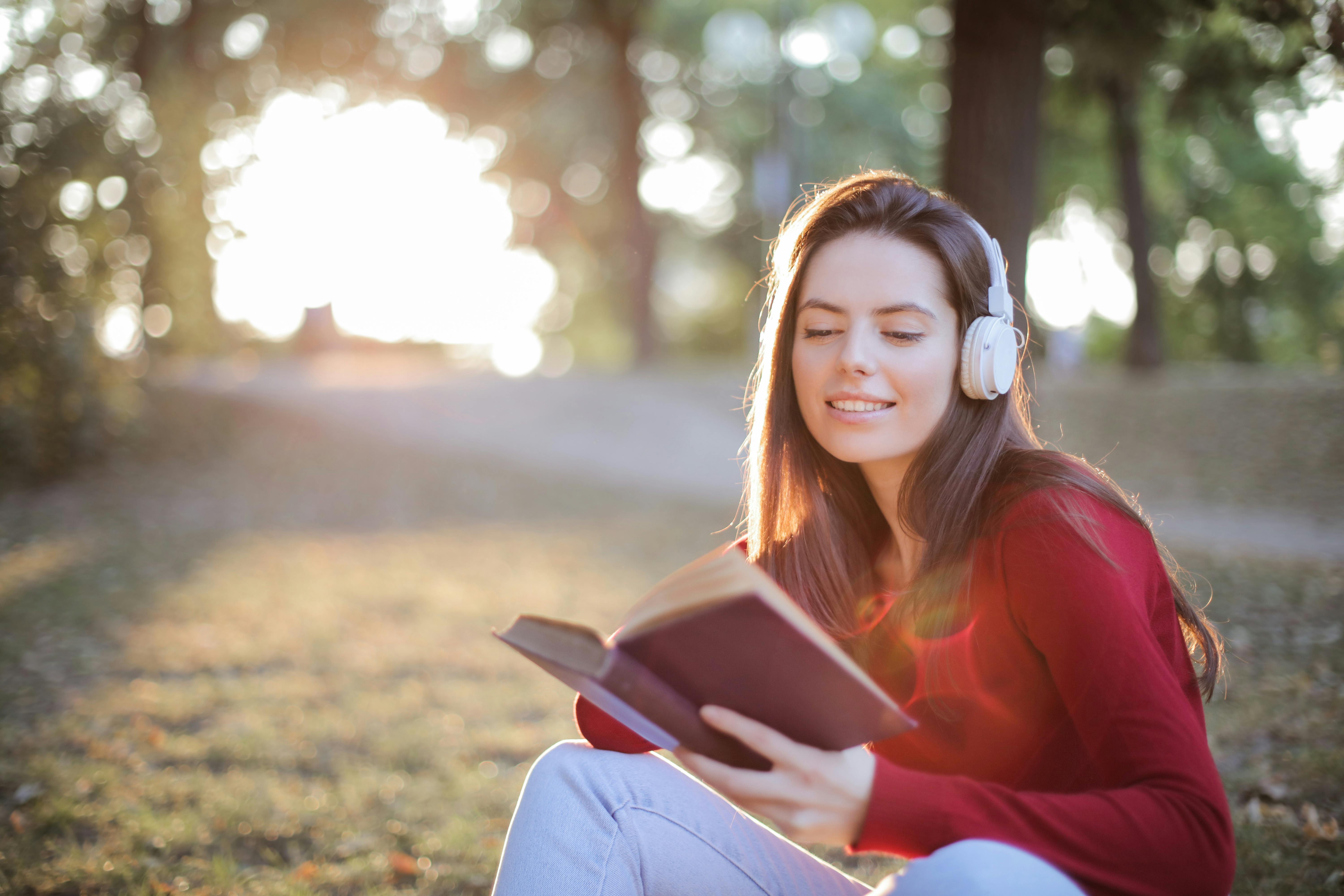 selective focus photo of smiling woman in a red long sleeve top reading book while listening to music on headphones