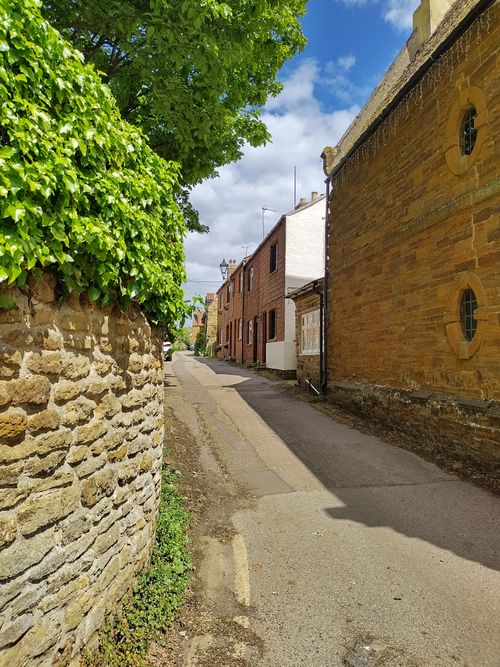 Free Sunlit village alley with rustic stone walls and greenery under a blue sky. Stock Photo