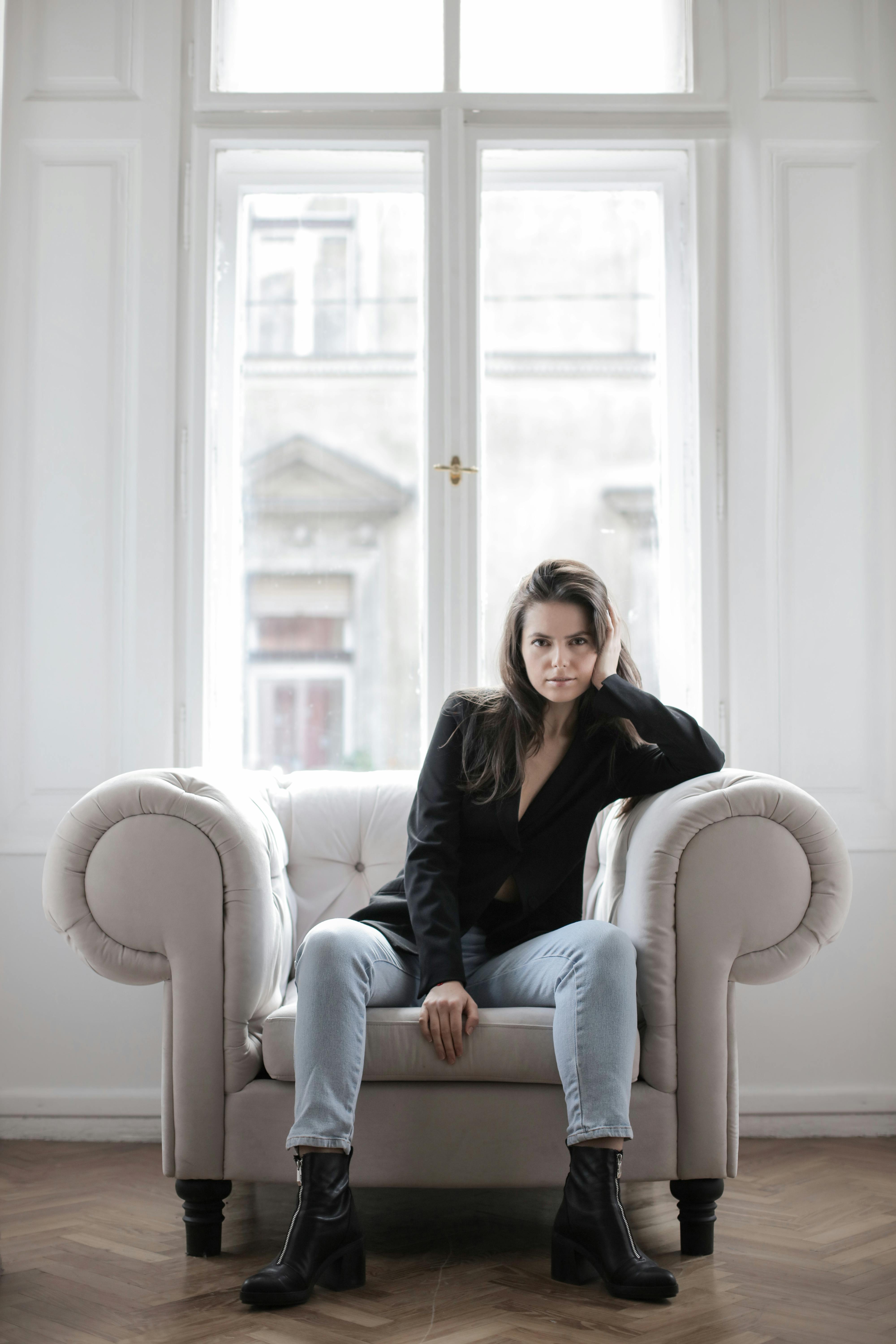 一位黑发女子穿着黑色外套和牛仔裤，坐在扶手椅上，窗外透进自然光线