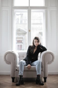 一位黑发女子穿着黑色外套和牛仔裤，坐在扶手椅上，窗外透进自然光线