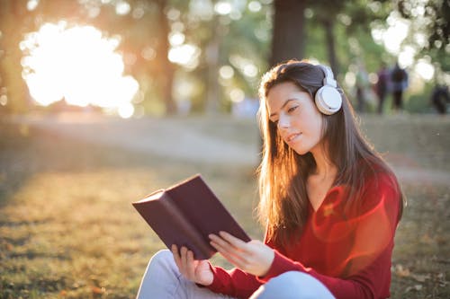 무료 헤드폰에서 음악을 들으면서 빨간색 긴 소매 상단 읽기 책에 웃는 여자의 선택적 초점 사진 스톡 사진