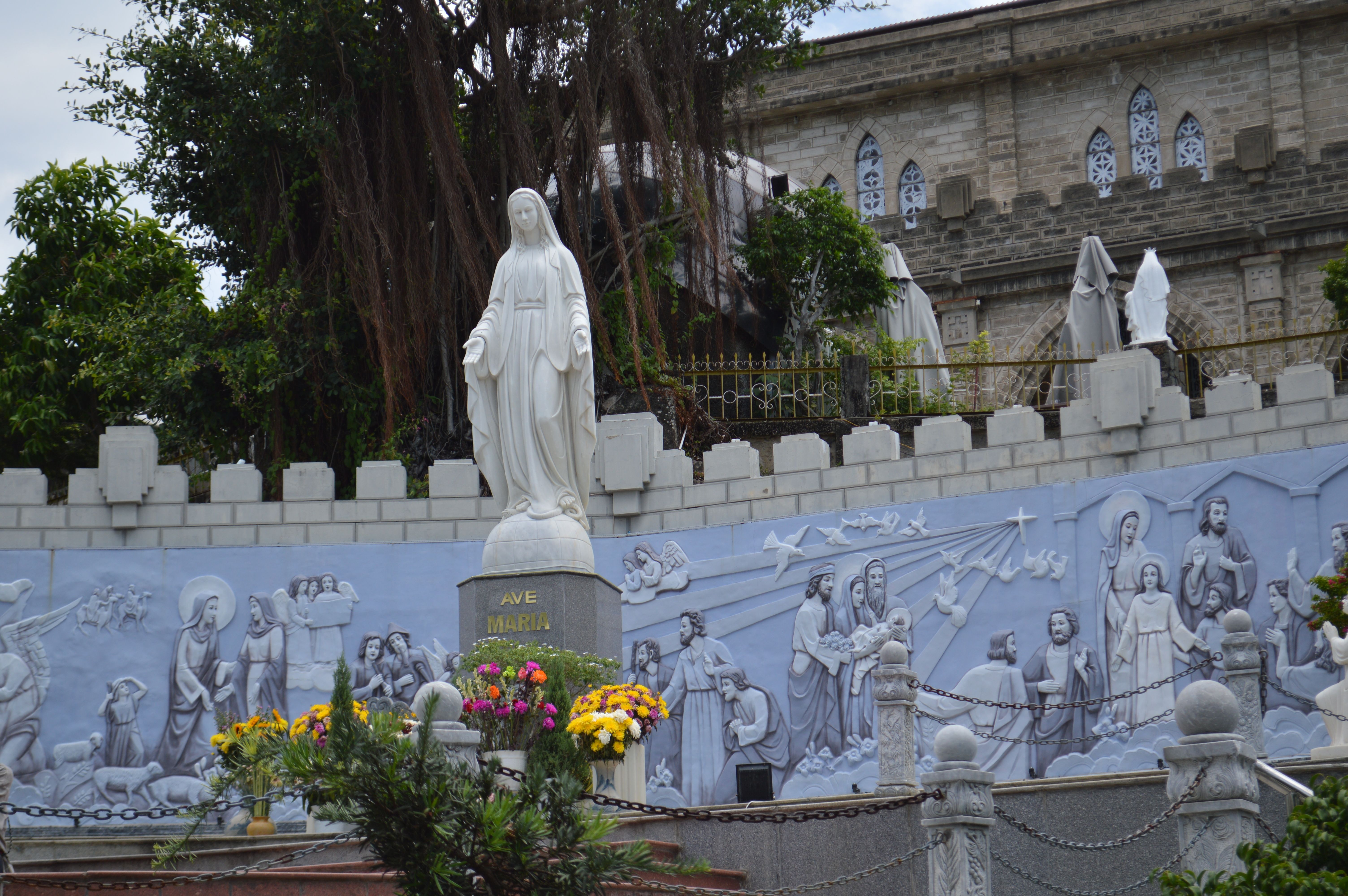 Outdoor statue of the Virgin Mary surrounded by floral arrangements, murals, and religious art.