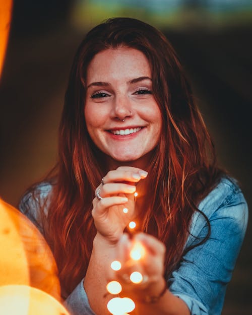 Free Selective Focus Photo of Smiling Woman in Denim Shirt Holding String Lights Stock Photo