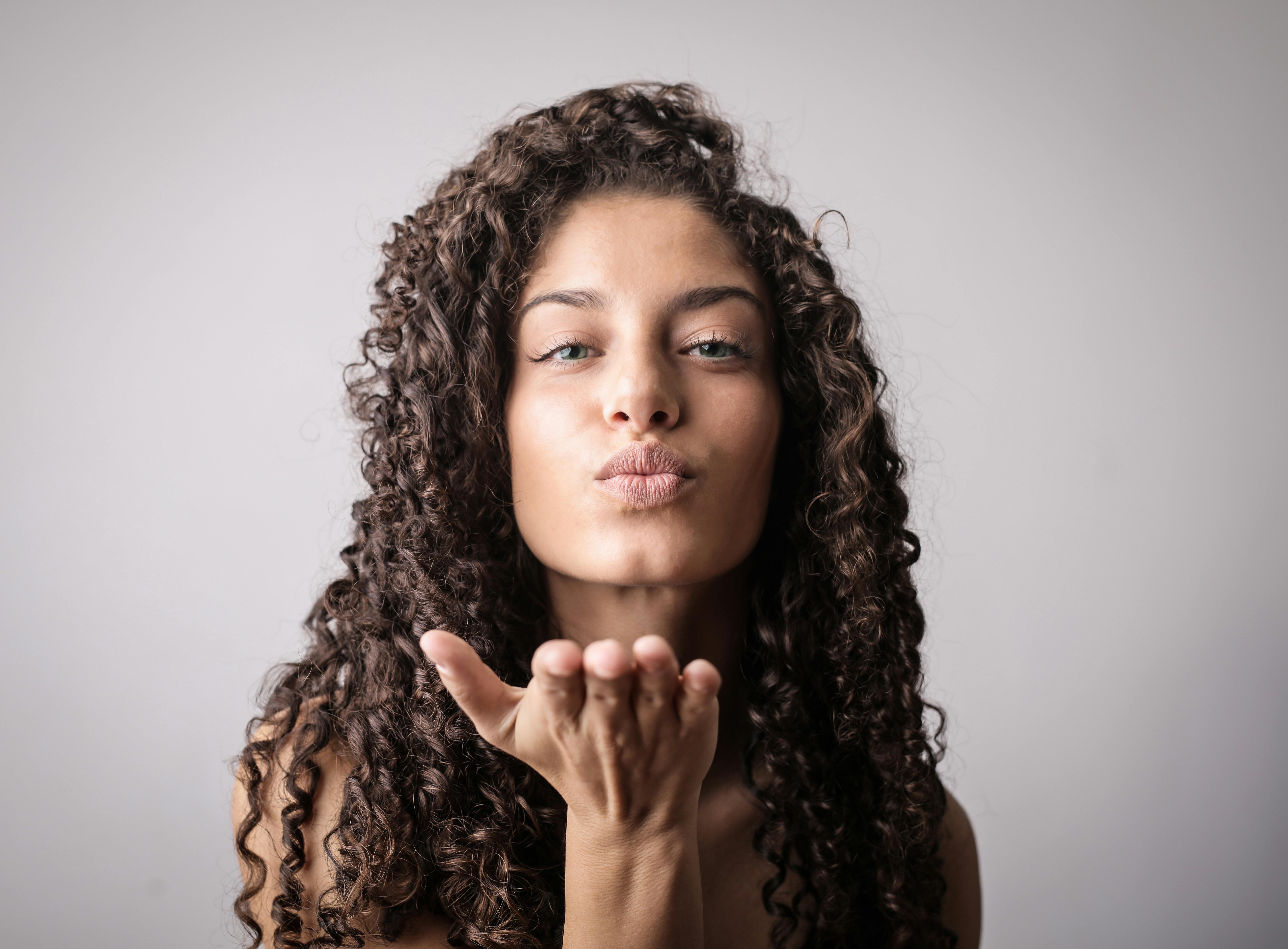 A woman with 3B curly hair blows a kiss towards the camera, exuding warmth and friendliness. She is set against a plain gray background, enhancing the focus on her gesture.