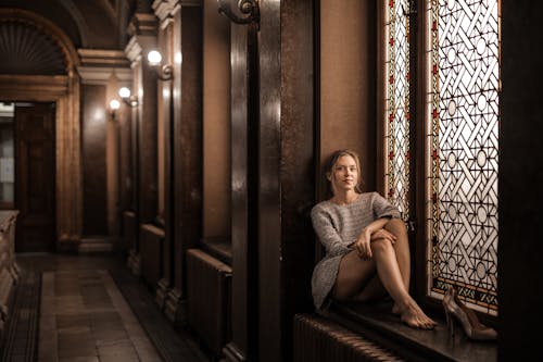 Blonde barefoot female sitting by painted window in dark lobby with Gothic interior