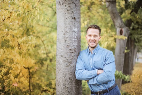 Man in Blue Dress Shirt Standing Beside Tree Trunks