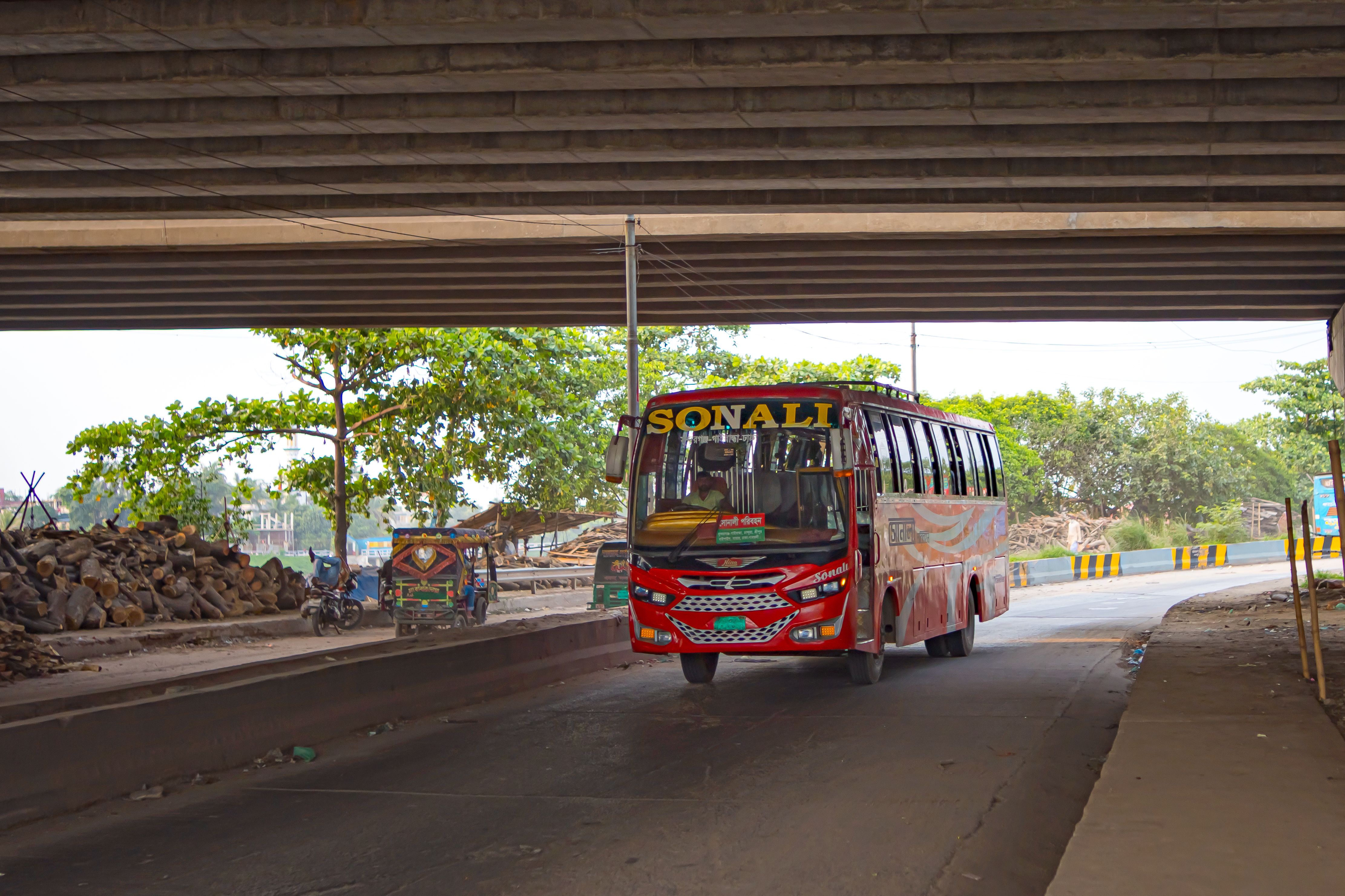 Red public bus driving under an urban overpass in Dhaka, Bangladesh, showcasing city life.
