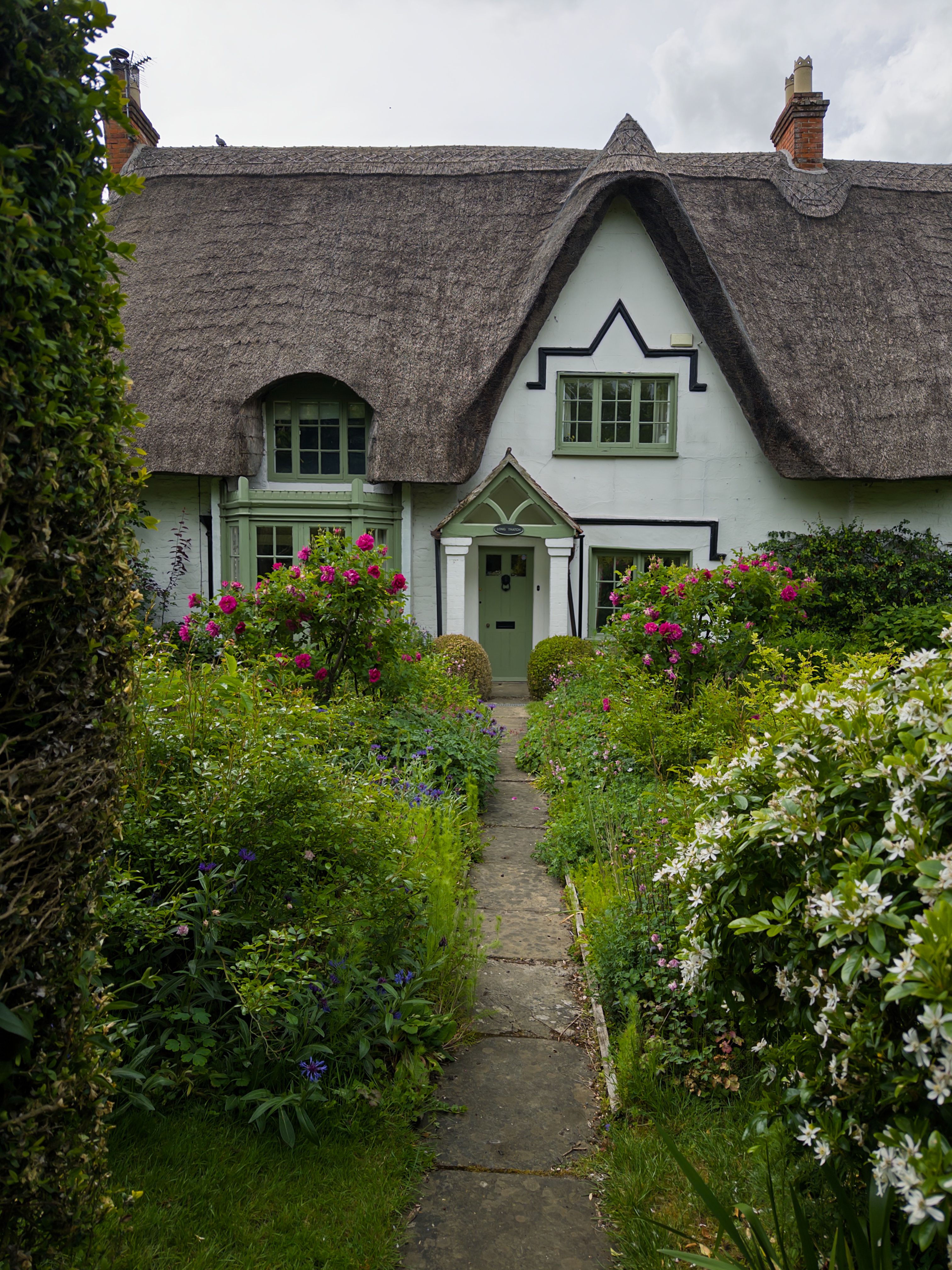 A picturesque white cottage with thatched roof surrounded by lush garden and blooming flowers in summer.