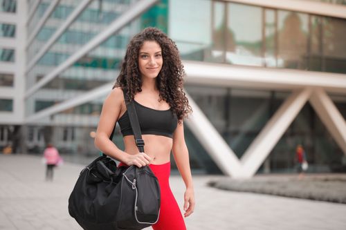 Free Young woman in activewear smiling with a gym bag outdoors in urban setting. Stock Photo