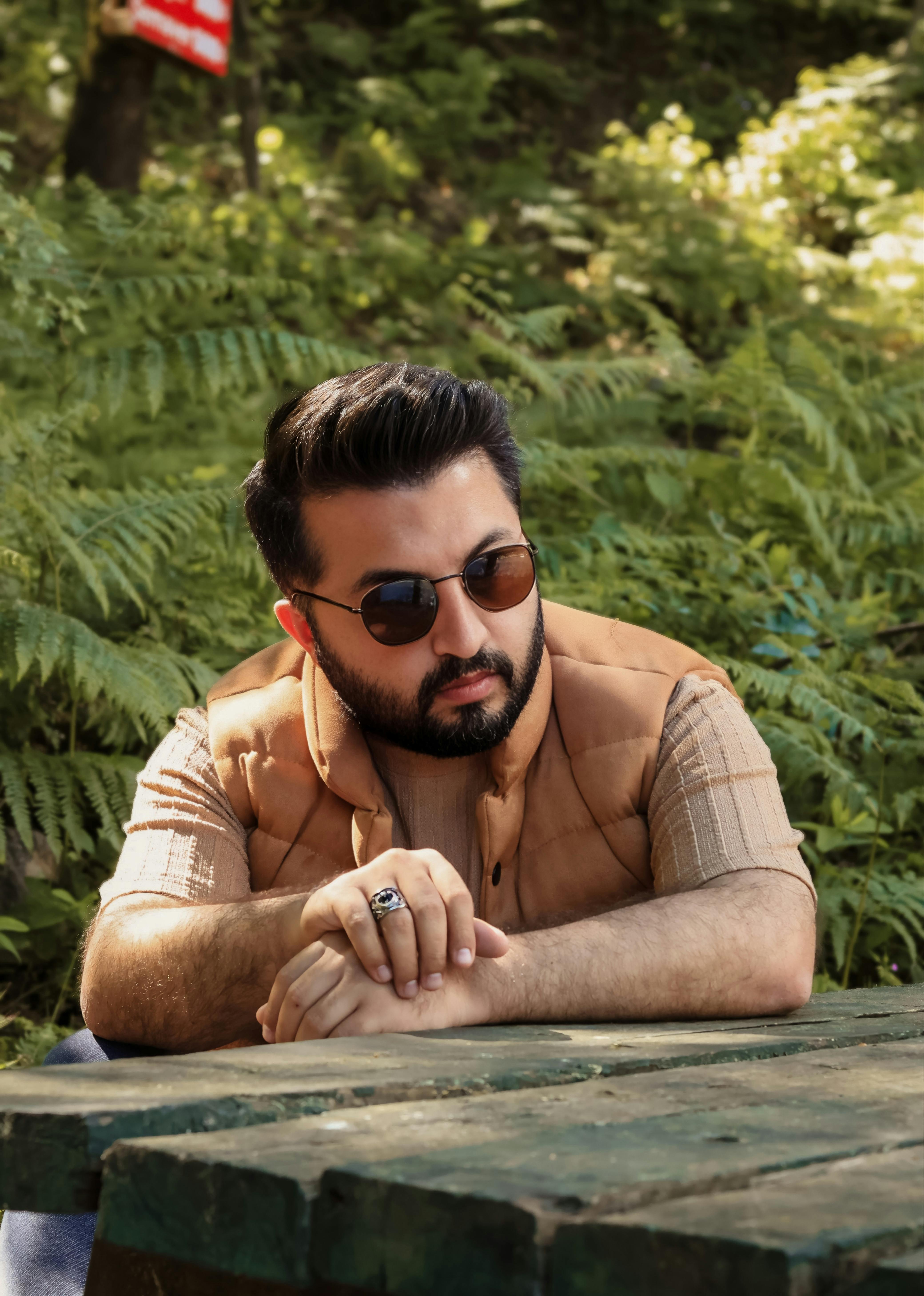 Man wearing sunglasses and vest, sitting casually at an outdoor picnic table in a lush forest setting.