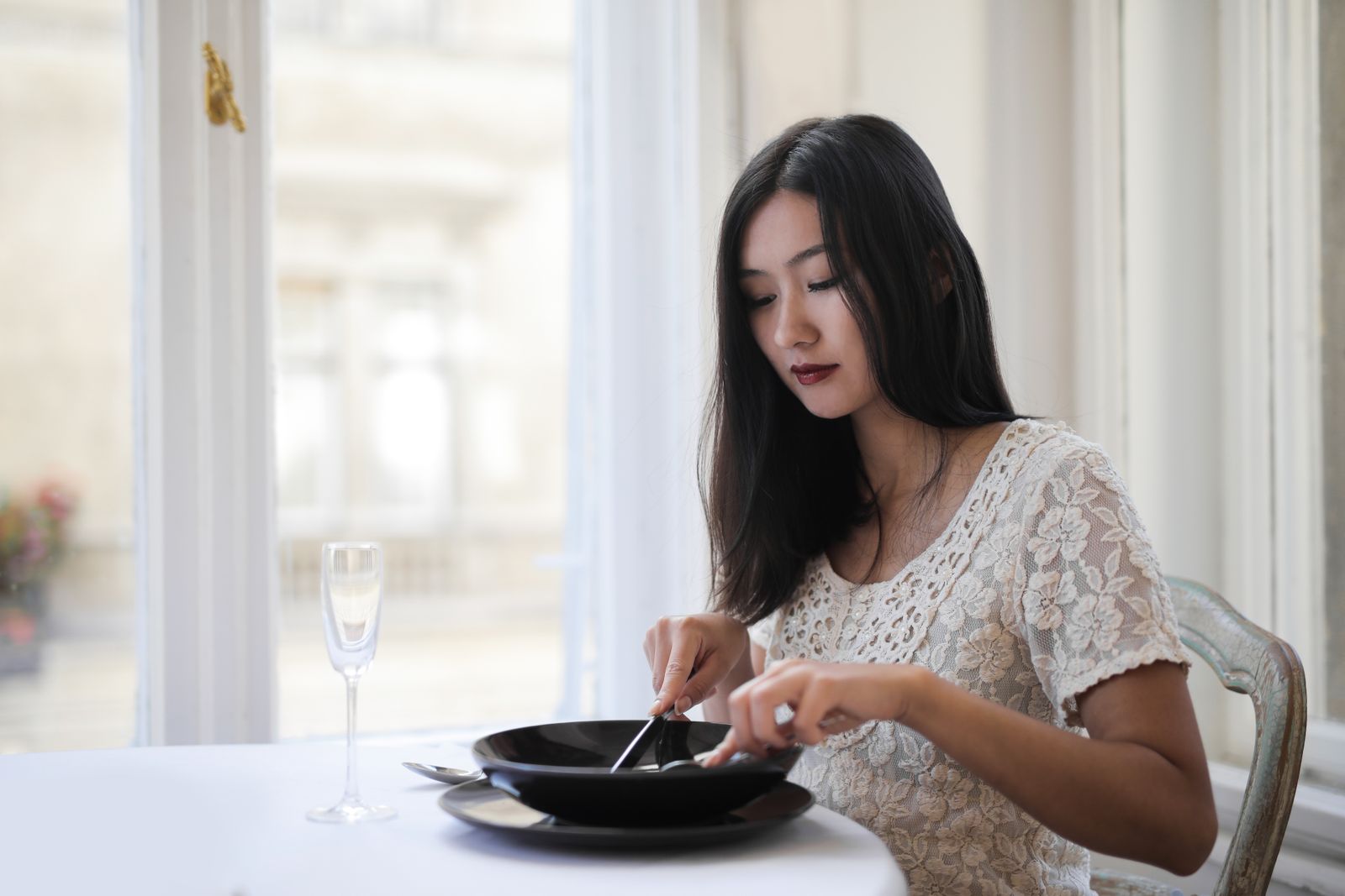 Woman with long dark hair in a lace top dining at a white table by a bright window