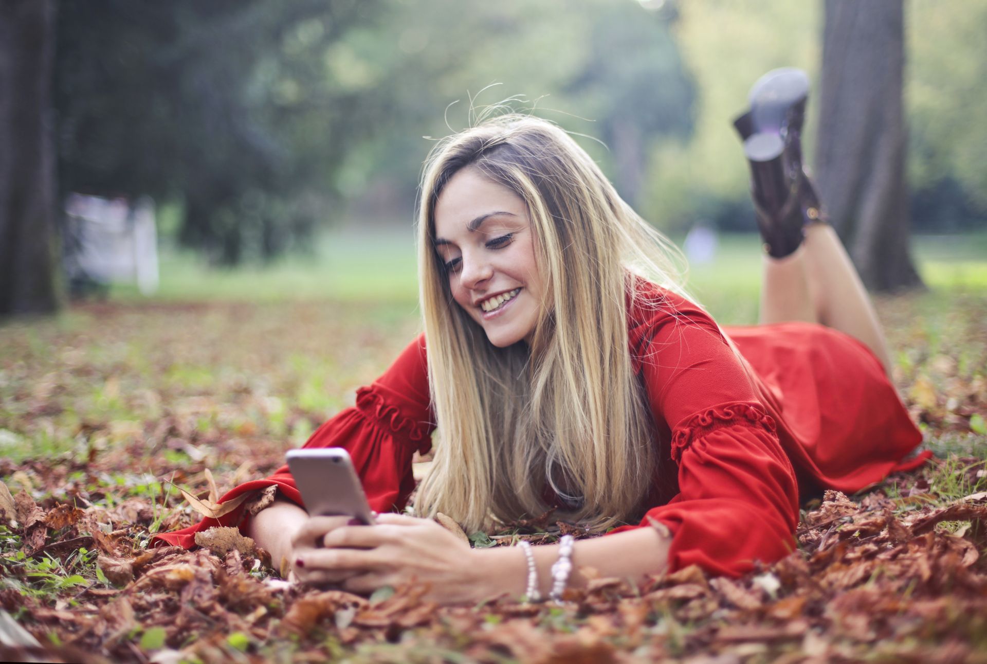 Smiling woman in red dress lying on autumn leaves texting on smartphone in a park.