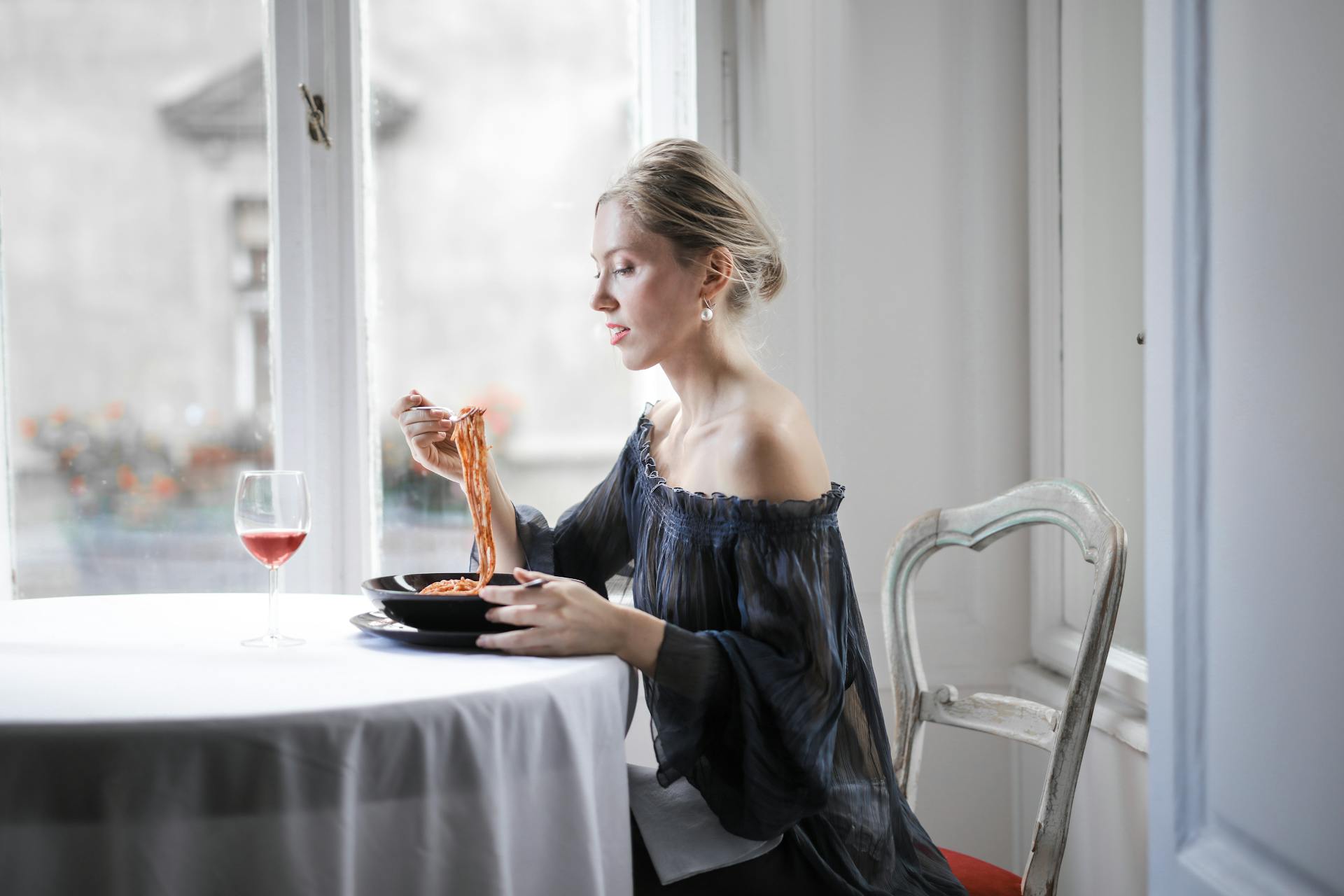 Selective Focus Photo of Woman in a Black Off Shoulder Dress Sitting at a Table Alone Eating Spaghetti