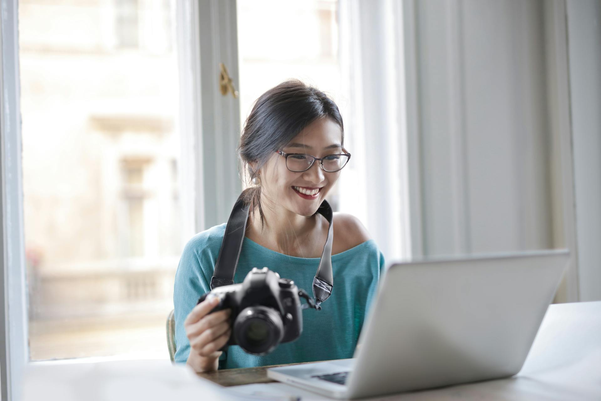 Happy Asian female with professional camera sitting at table with laptop in cozy room