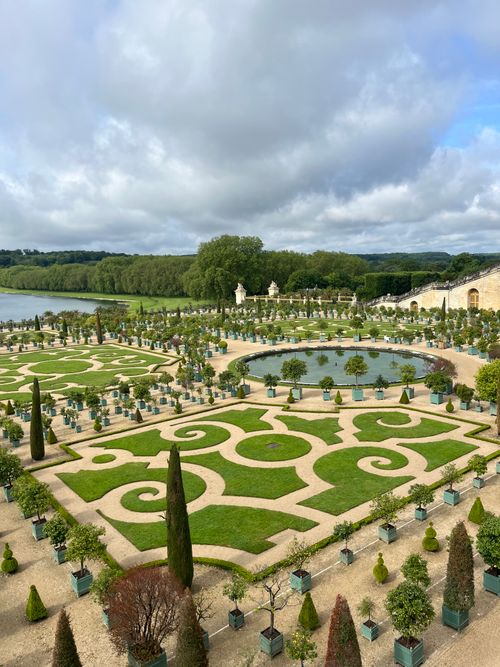 Free Explore the intricate ornamental gardens of Versailles, set against a lush landscape and cloudy sky. Stock Photo