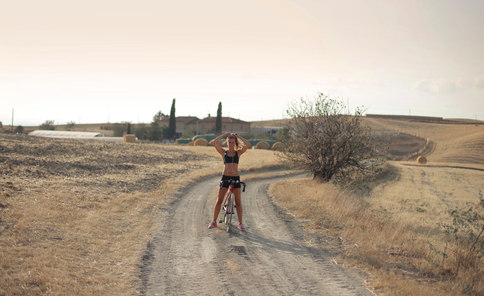 Young female in sportswear standing with cycle on road in remote countryside while doing ponytail