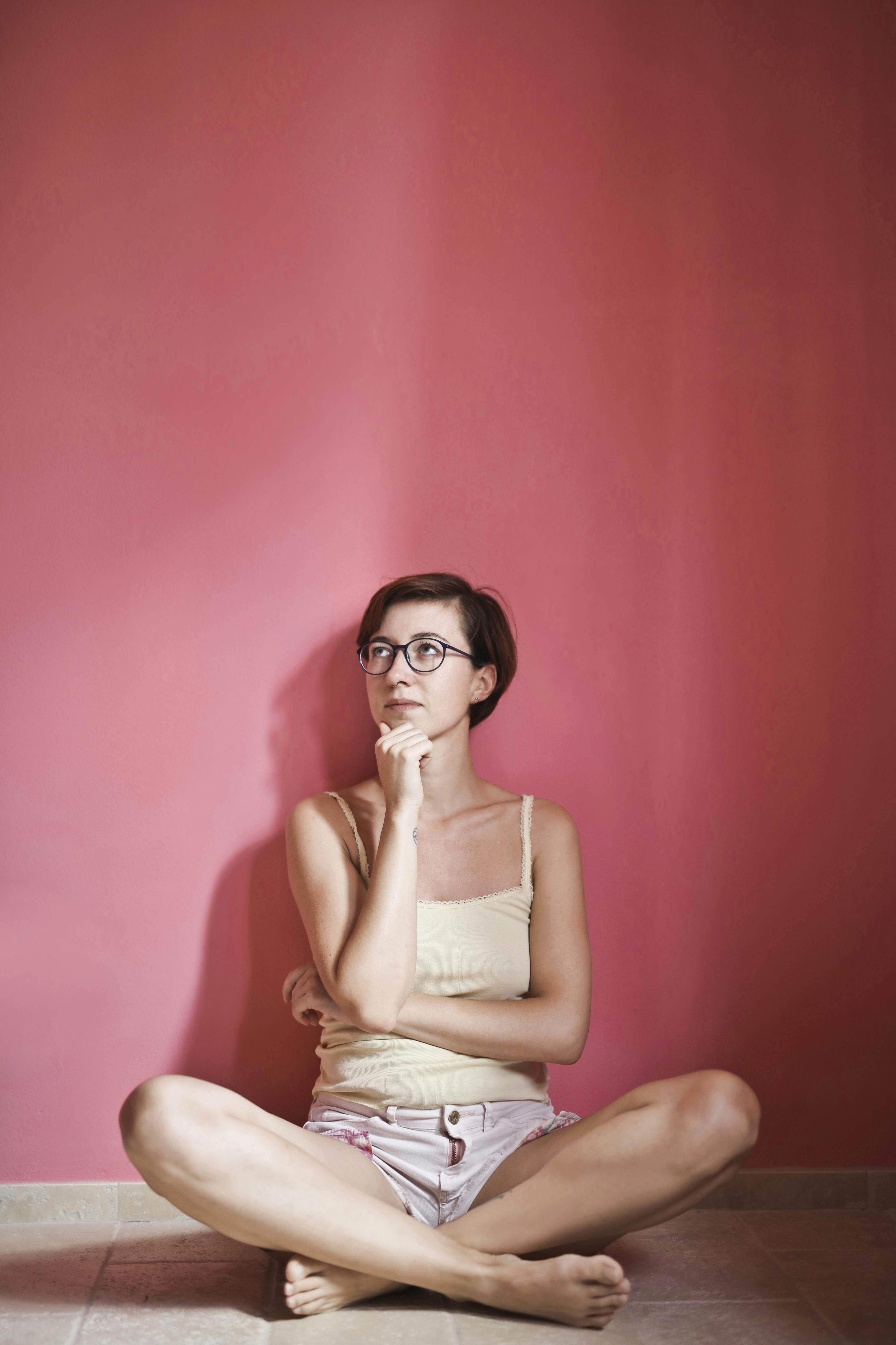 photo of thinking woman in yellow tank top and pink shorts sitting on floor next to pink wall