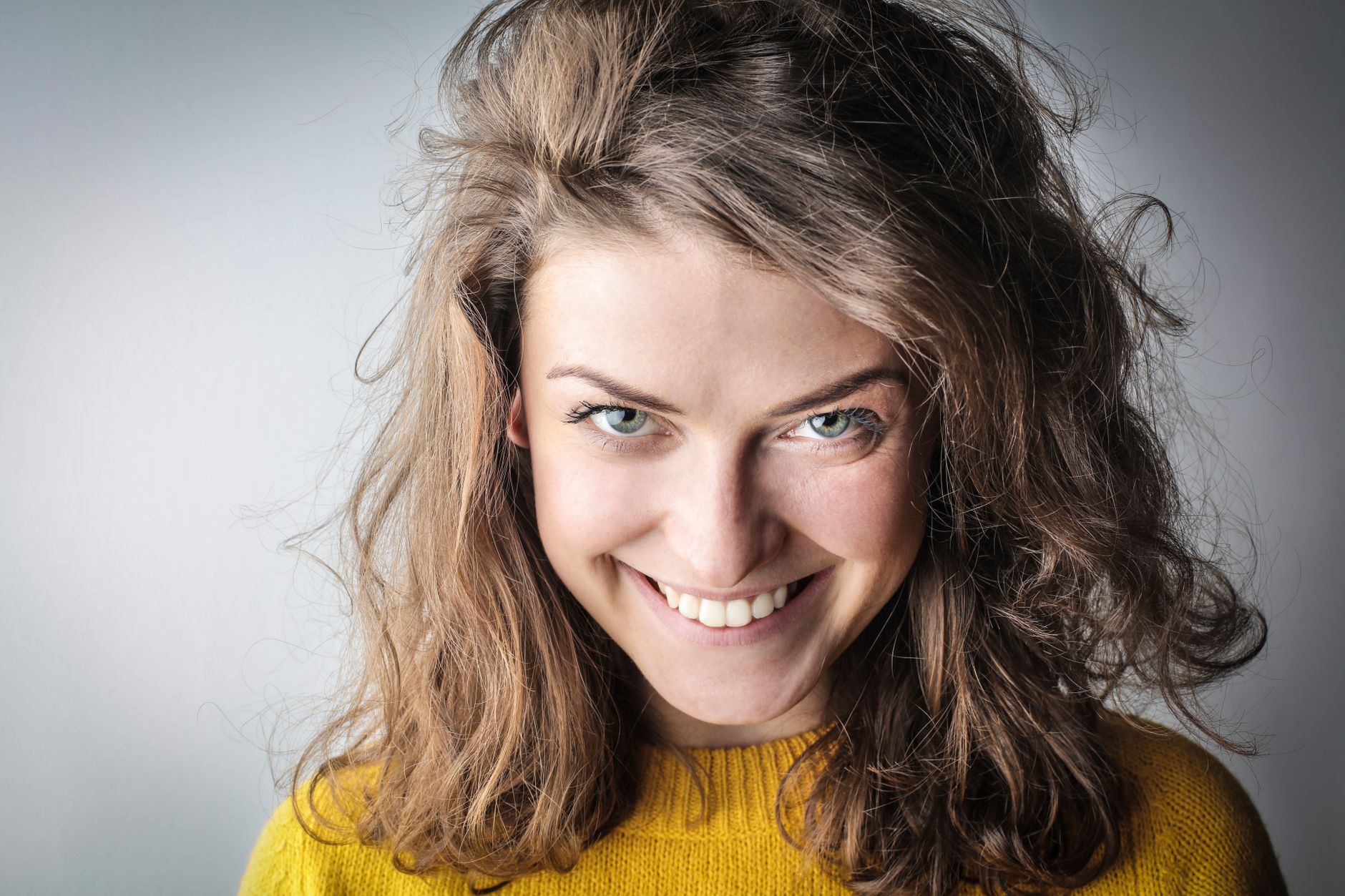Close-up portrait of a smiling woman in a yellow sweater and casual hair, exuding happiness against a gray background.