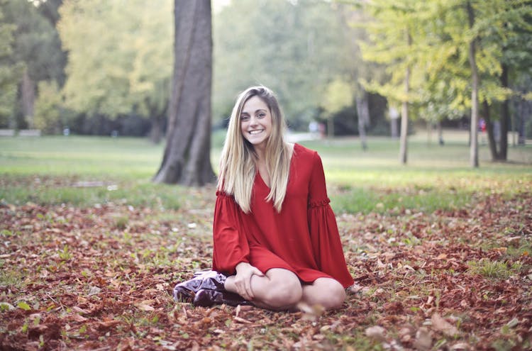 Photo Of Smiling Woman In Red Long Sleeve Dress Sitting Alone On Brown Dried Leaves