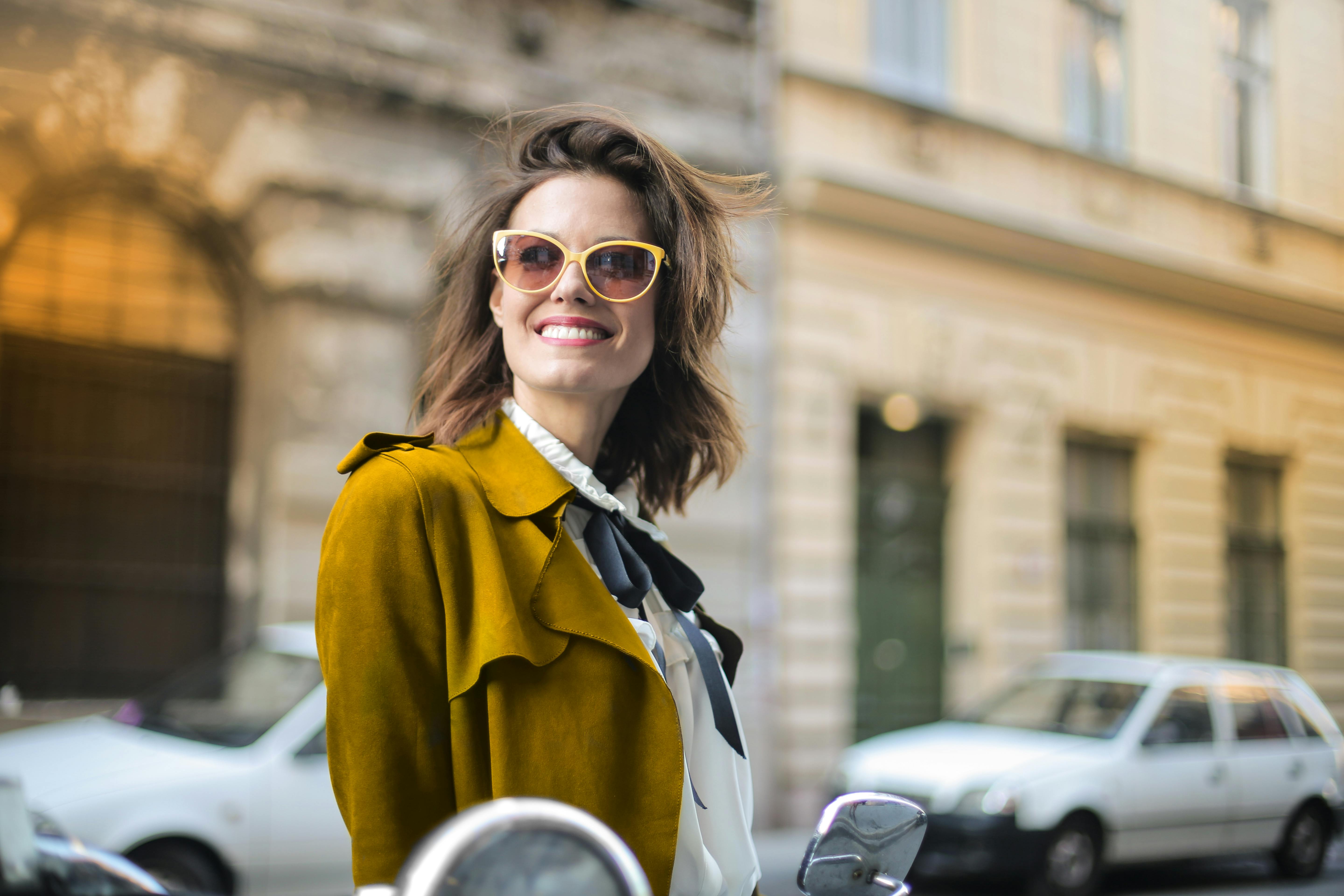 一位戴着太阳镜的女士，穿着别致的黄色外套，在城市街道上自信地微笑着
