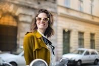 一位戴着太阳镜的女士，穿着别致的黄色外套，在城市街道上自信地微笑着