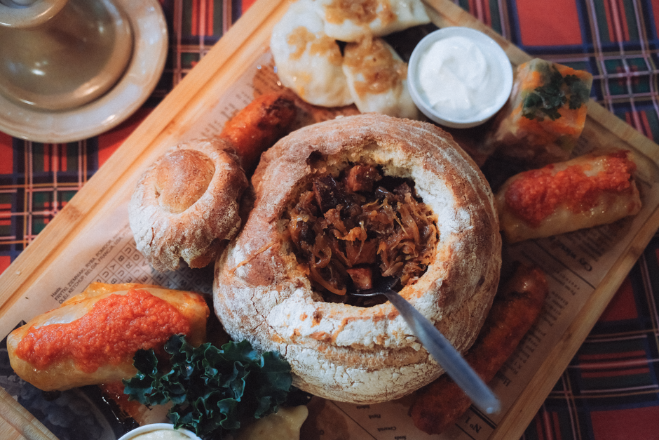 A wooden table set with pierogi, sliced kielbasa, a bowl of bigos stew, rye bread, and a dish of sauerkraut.