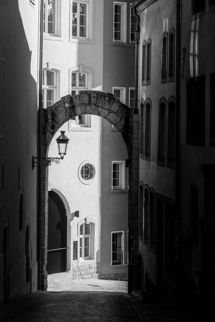 Dramatic black and white narrow European street with historic archway in sunlight and shadows.