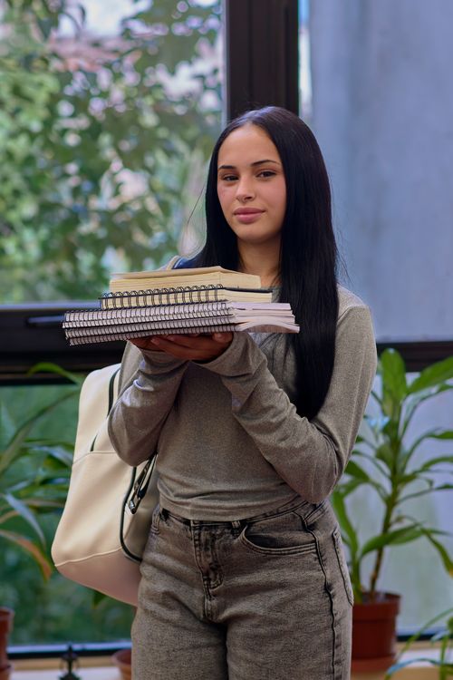 Free Young woman indoors holding a stack of books, wearing casual clothing. Stock Photo