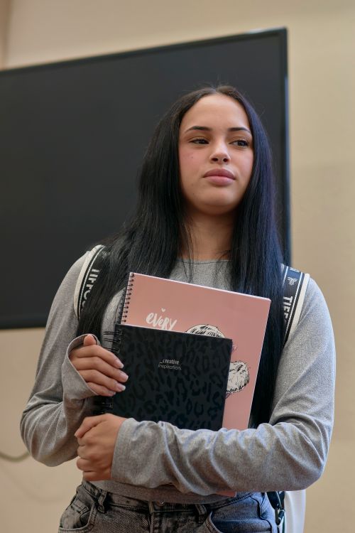Free A young woman standing indoors, holding notebooks and wearing a backpack, conveys a casual academic setting. Stock Photo