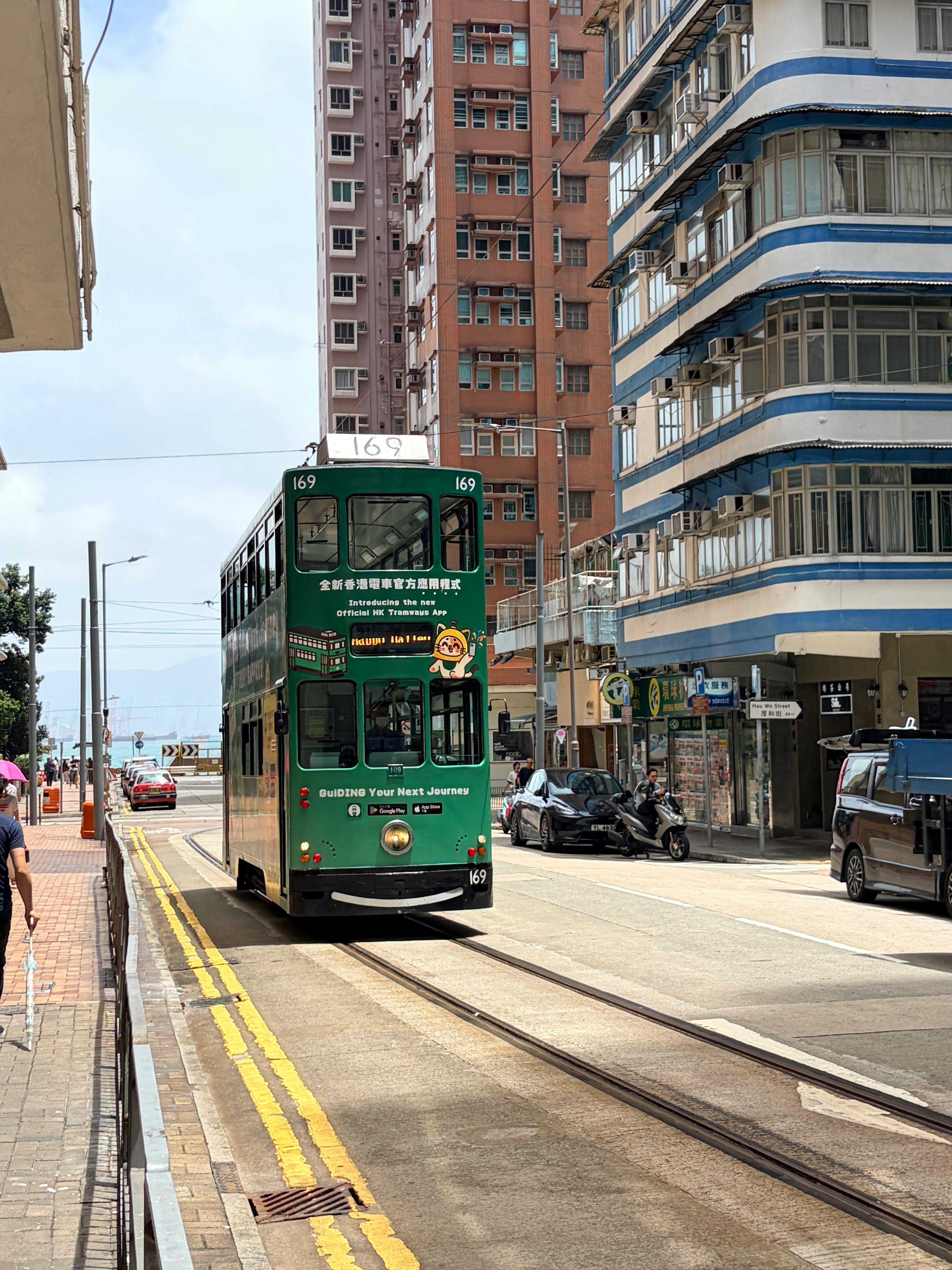 A classic green double-decker tram travels through bustling Hong Kong street on a sunny day.