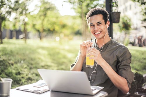 Cheerful guy with laptop and earphones sitting in park while drinking juice and smiling at camera
